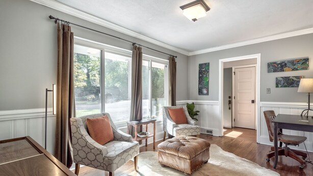 A spacious living room with two armchairs, a coffee table, and a leather ottoman. There are large windows with curtains, a door, and a painting on the wall. - Caught in the City-Walk to BoPlex Coliseum and Ovens Auditorium (Charlotte)