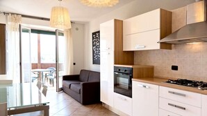 Eine moderne Küche mit weißen Schränken, einem Gasherd und einem Einbauofen. Es gibt eine Essecke mit einem Glastisch und Stühlen, und ein Sofa im Hintergrund.