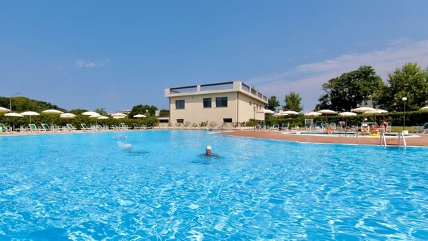 Ein Schwimmbecken mit Liegestühlen und Sonnenschirmen, im Hintergrund ein Gebäude und ein klarer Himmel.