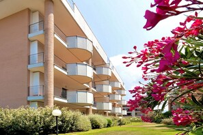 Un immeuble à plusieurs étages, doté de balcons et agrémenté d’un jardin bien entretenu où fleurissent des plantes aux fleurs roses.