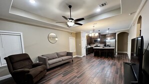 Un salón amplio con piso de madera oscura, ventilador de techo, sofá, sillón y televisor.