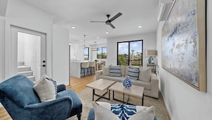 A modern living room with a sofa, armchair, and coffee table, a dining area with chairs, and a view of a garden through large windows. - Rooftop Ocean-View Home, Private Parking, Near Beach (Oceanside)
