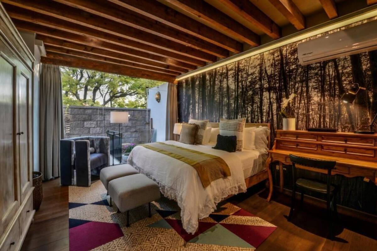 Un dormitorio con techo de madera, una cama con cubrecama blanco, un banco, un escritorio y una pared de piedra.