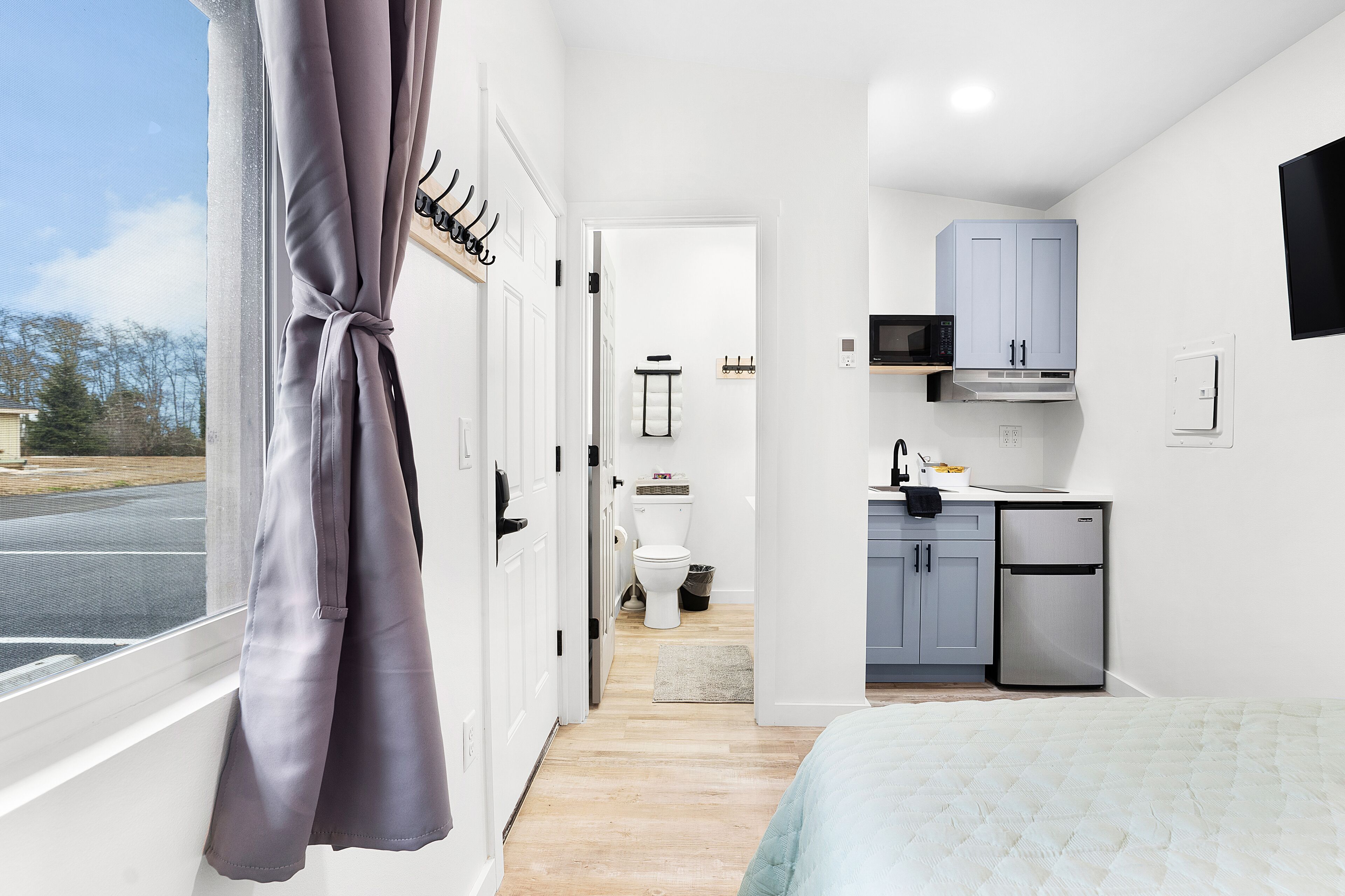 A compact bedroom with a bed, a window with curtains, a view of the outside, and a kitchenette with a microwave, cabinets, and a refrigerator.