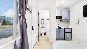 A compact bedroom with a bed, a window with curtains, a view of the outside, and a kitchenette with a microwave, cabinets, and a refrigerator.