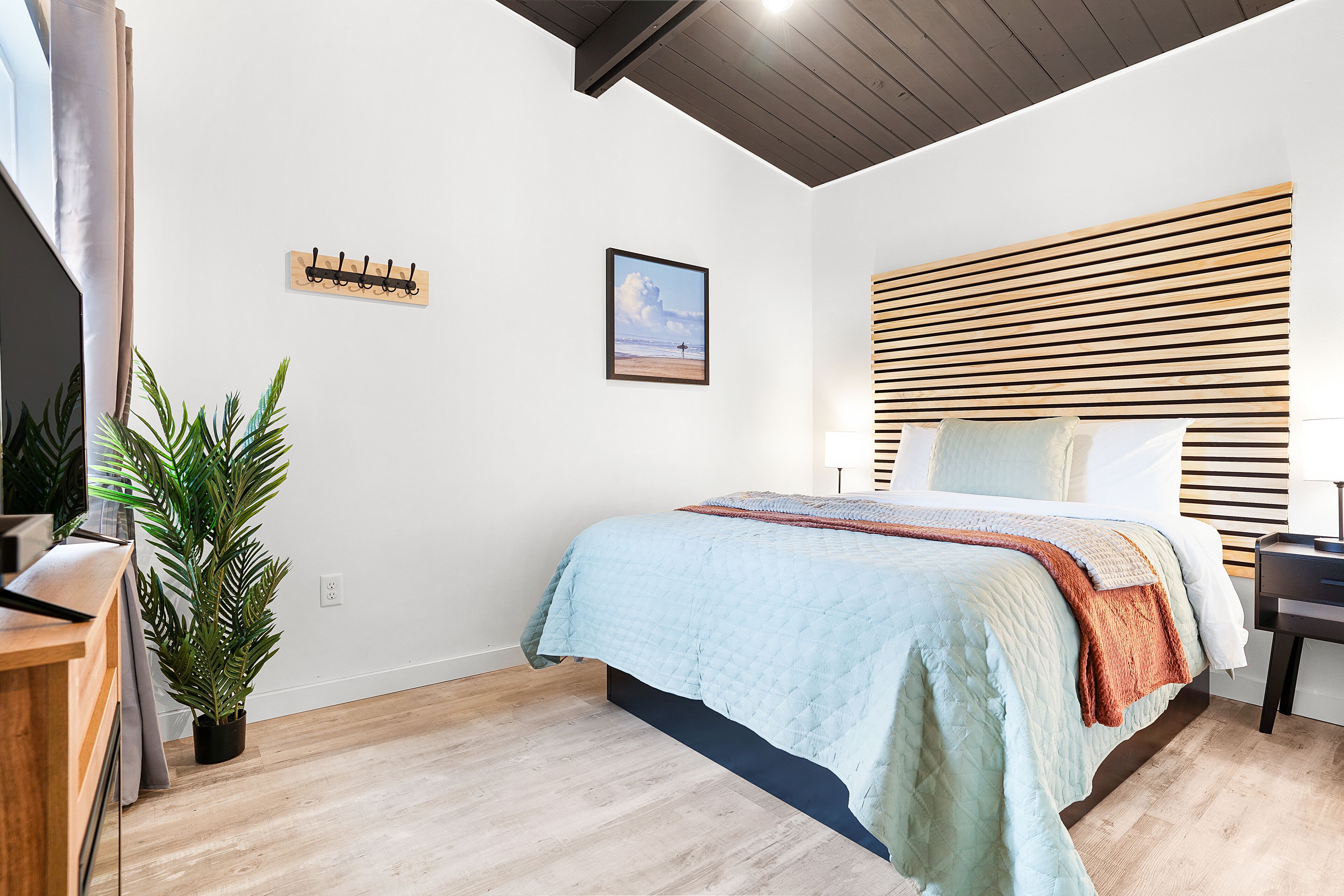 A bedroom with a bed, a wooden headboard, a plant, a coat rack, and a framed picture on the wall.