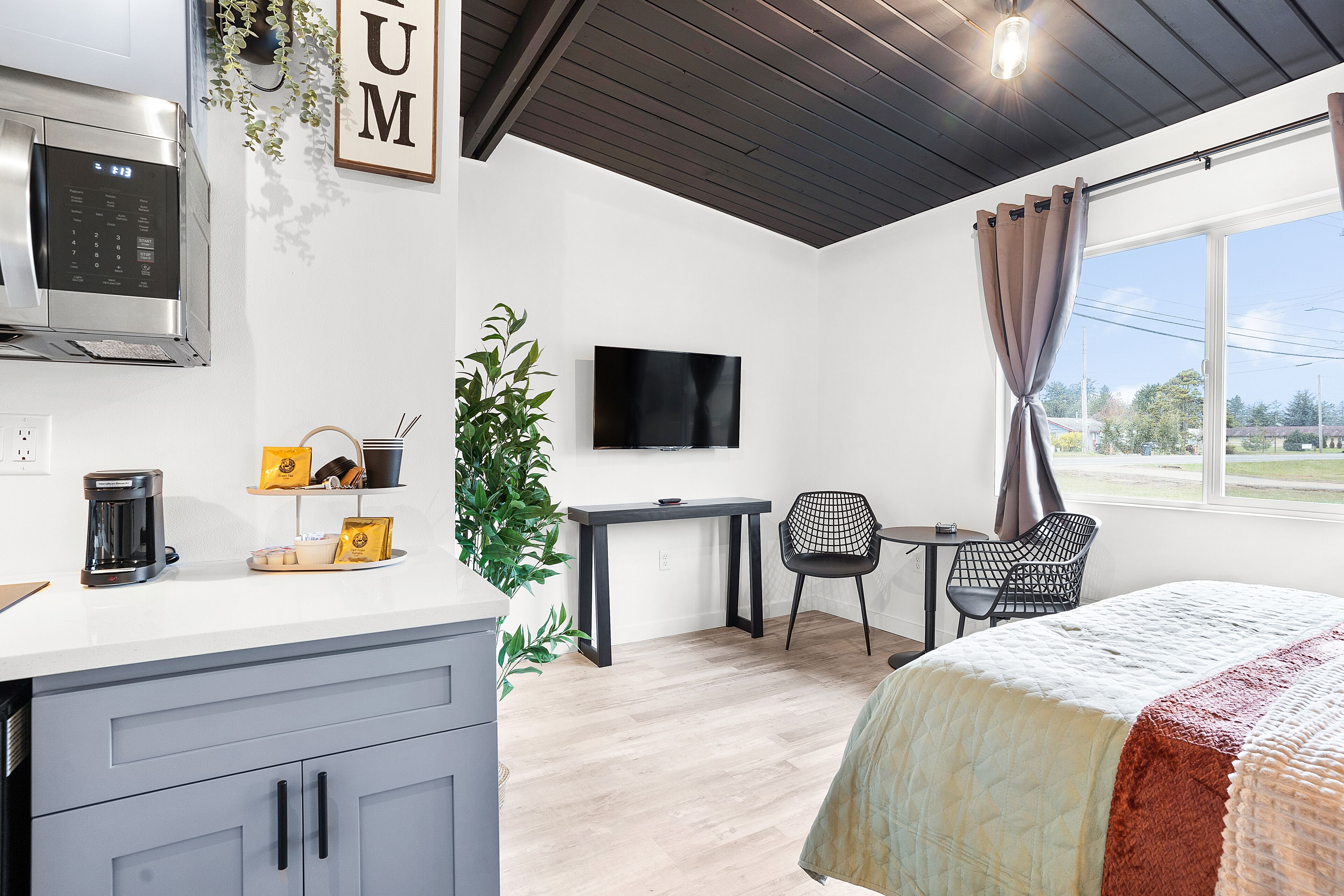 Quarto de hotel moderno com cozinha compacta, televisão de tela plana, cama com colcha, escrivaninha e uma cadeira.