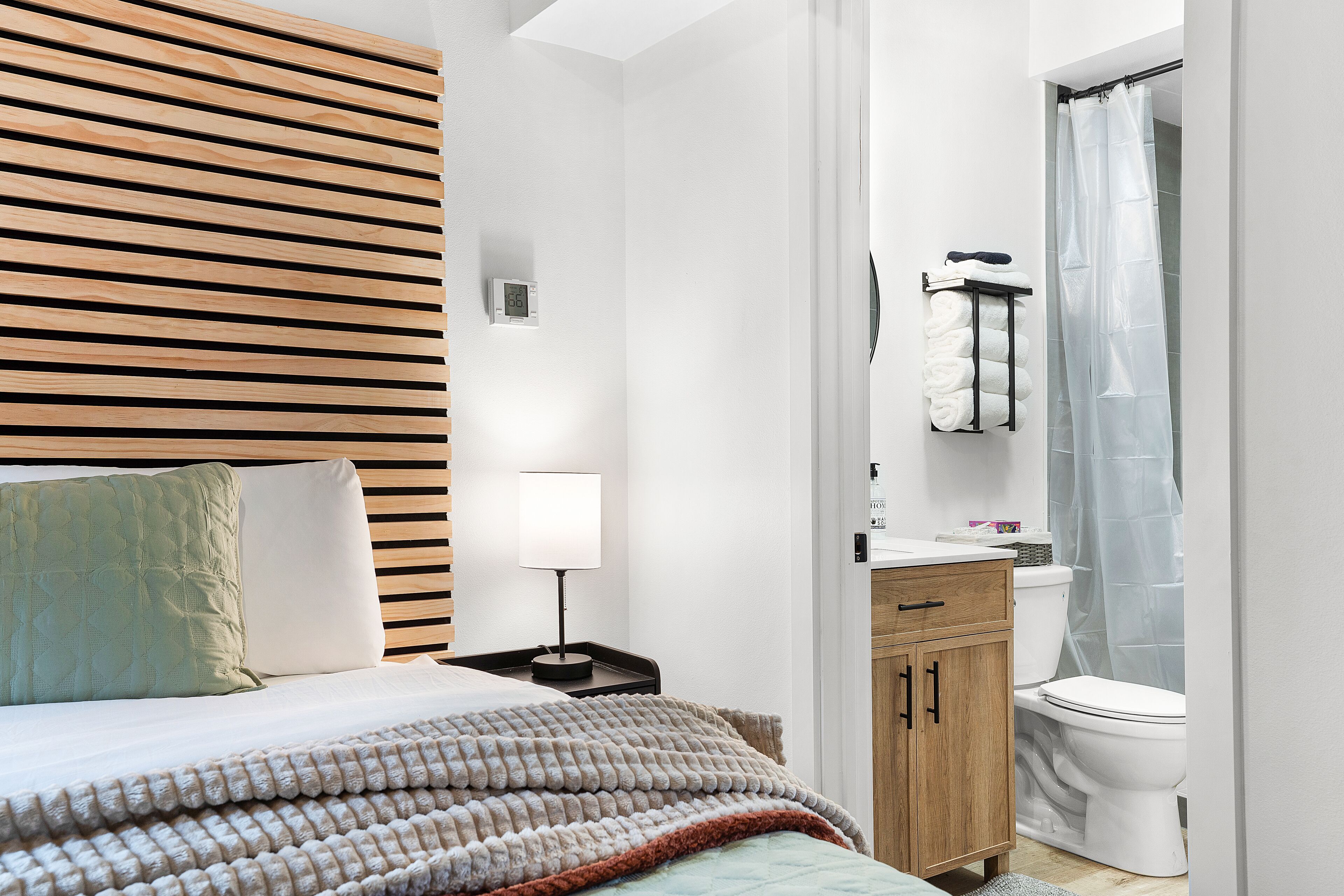 A modern bedroom with a wooden headboard, a bed with a quilted blanket, a bedside table with a lamp, and a wooden cabinet.