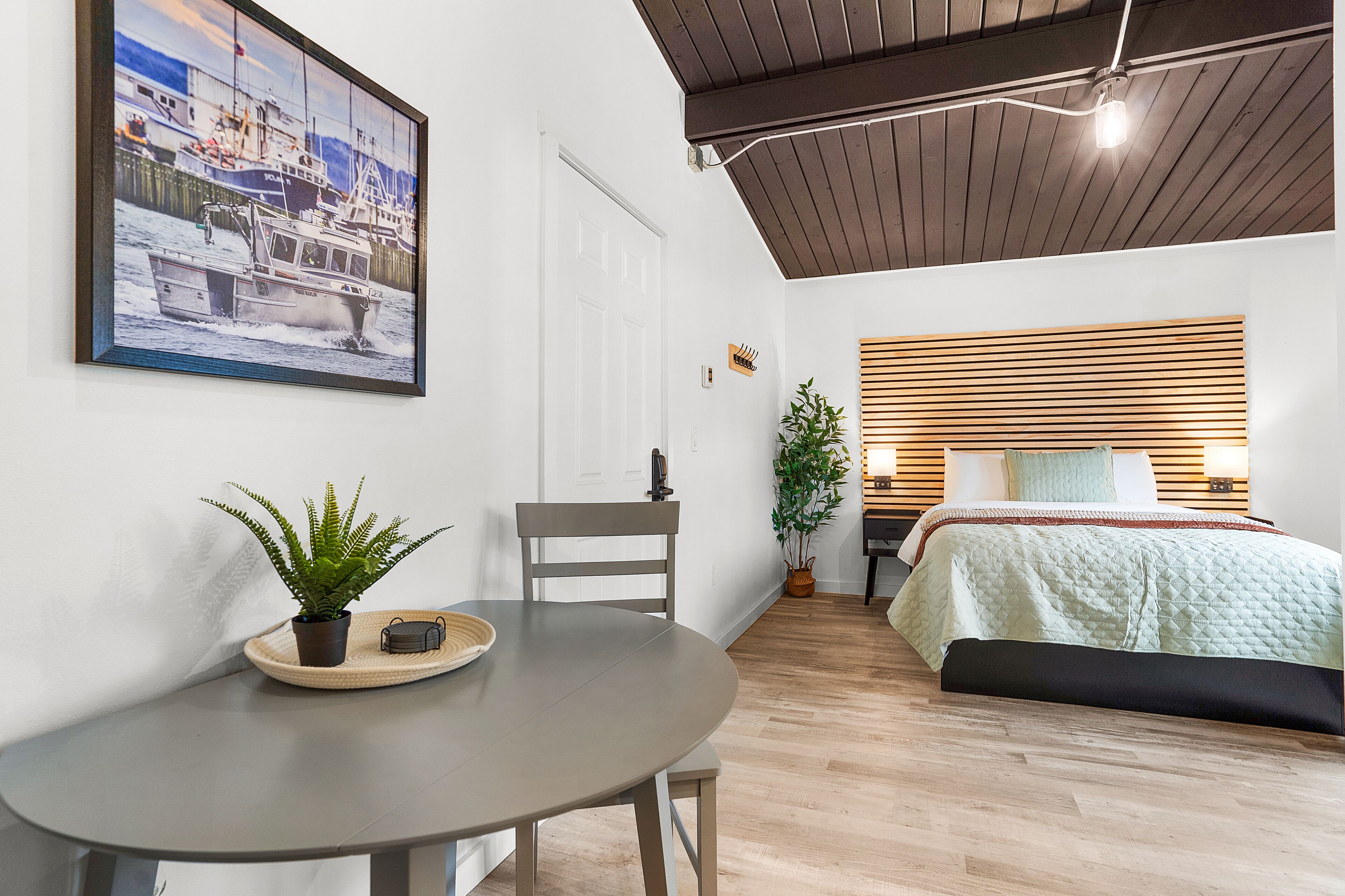 A room with a bed, a dining table with a plant, a framed picture on the wall, and a wooden ceiling.