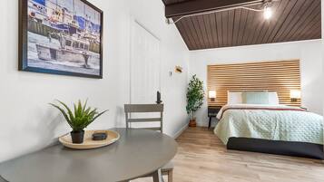 A room with a bed, a dining table with a plant, a framed picture on the wall, and a wooden ceiling.