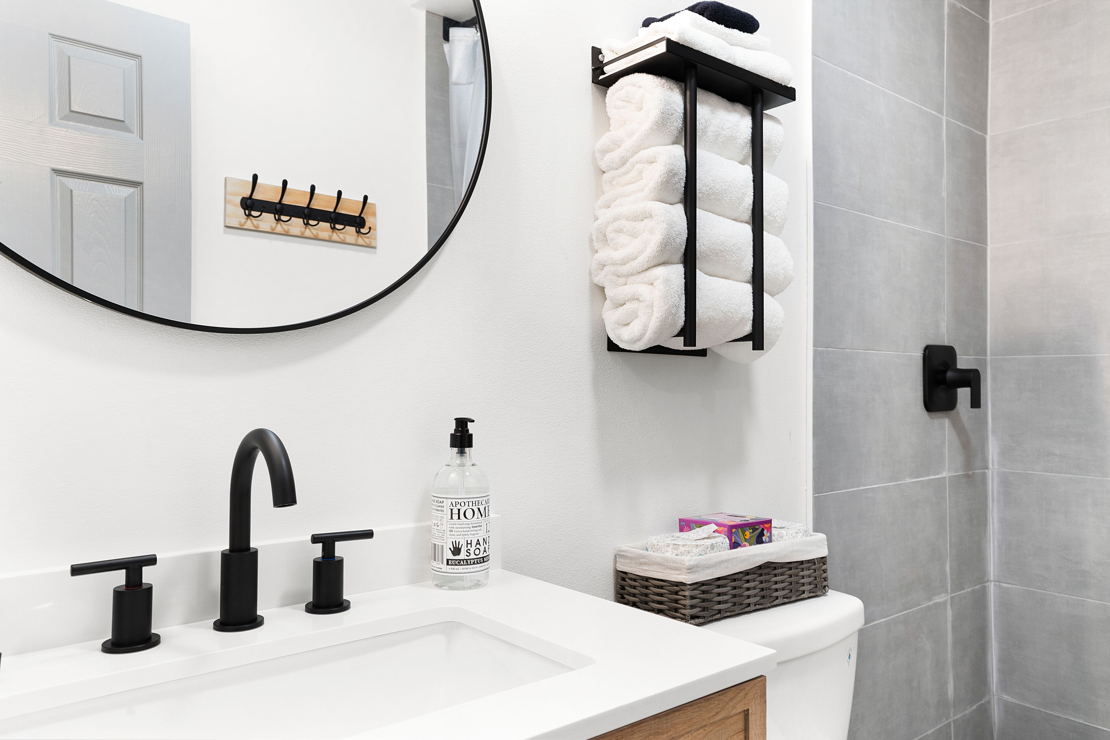 A bathroom with a white sink, black faucets, a round mirror, a towel rack with folded towels, a soap dispenser, and a wicker basket with toiletries.