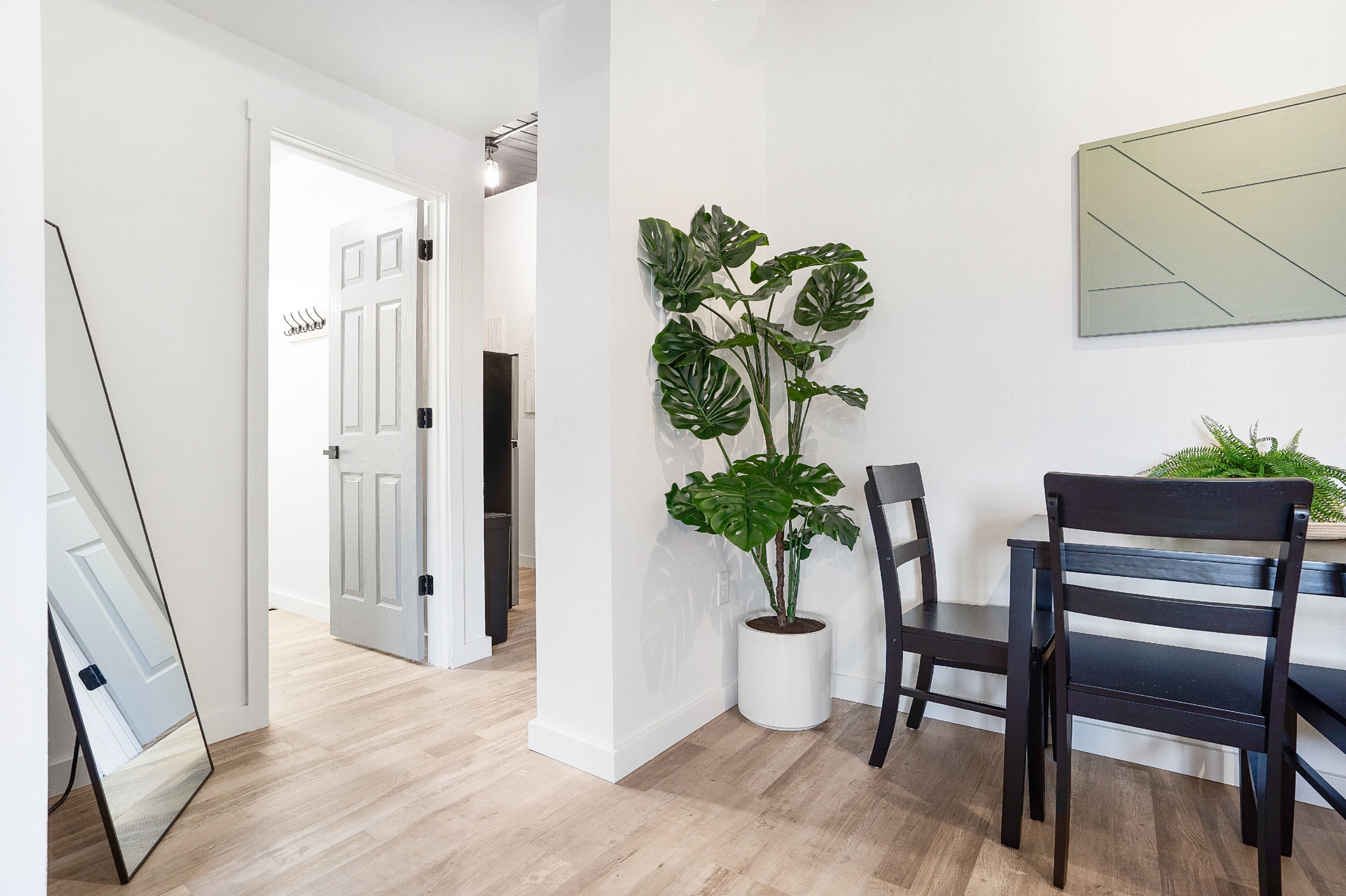 A modern interior with a wooden floor, a potted monstera plant, a dining table with chairs, and a large mirror.