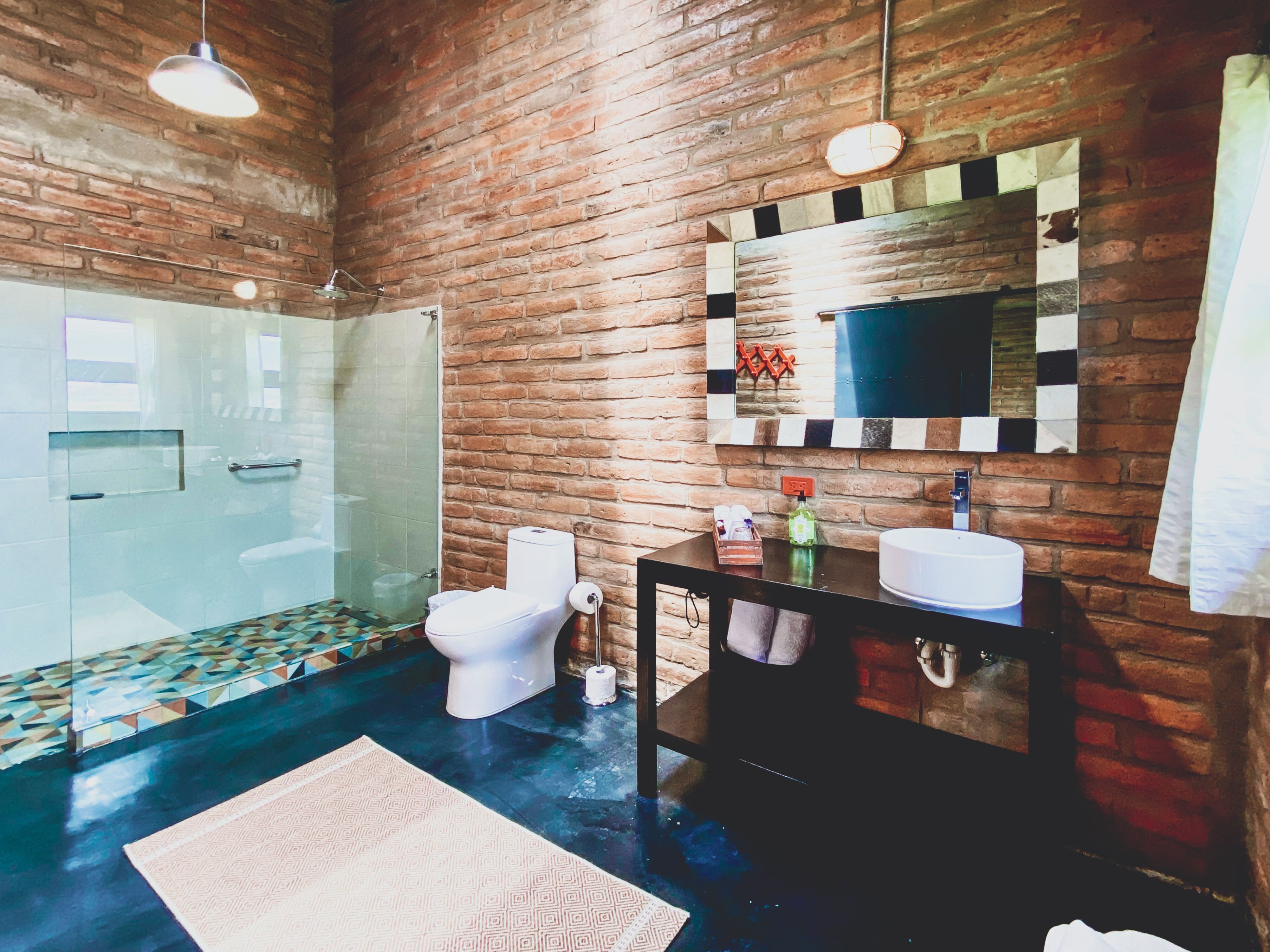 A bathroom with a glass shower, a toilet, a sink, and a mirror on a brick wall.