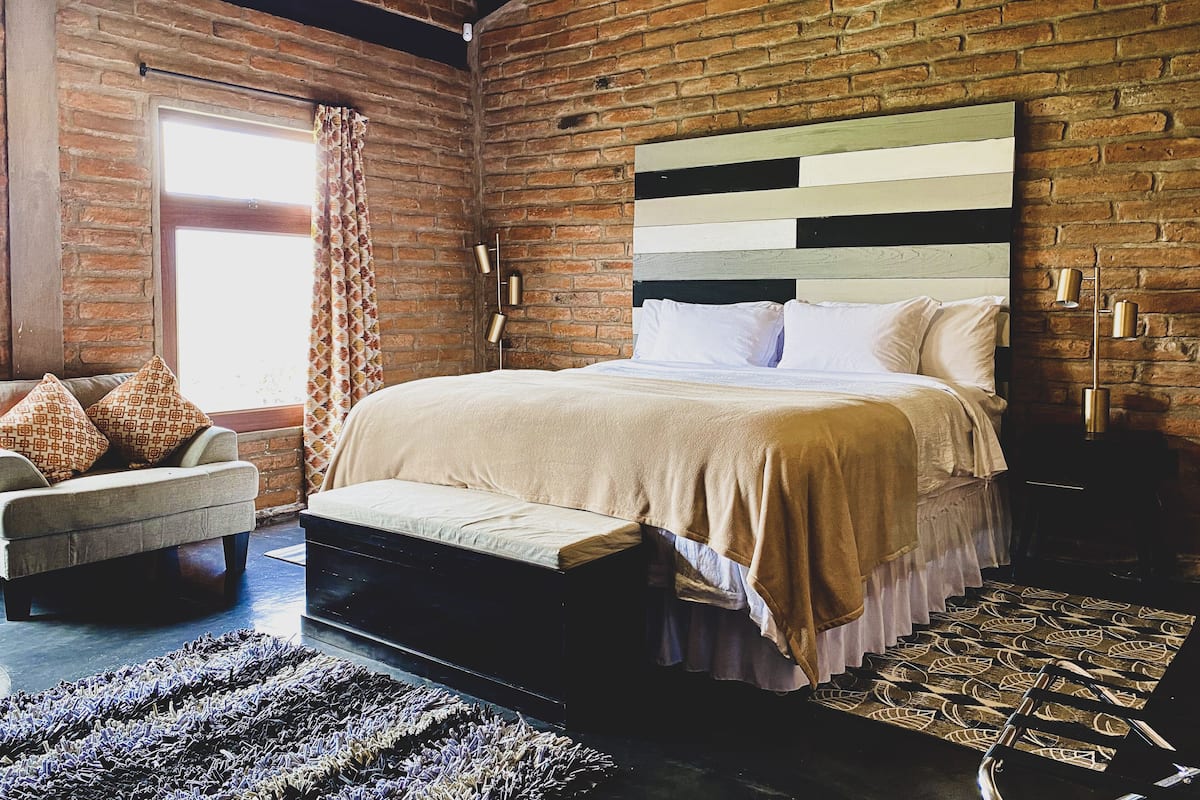 Un dormitorio con pared de ladrillo, una cama con cabecera a rayas, un banco y una alfombra estampada.