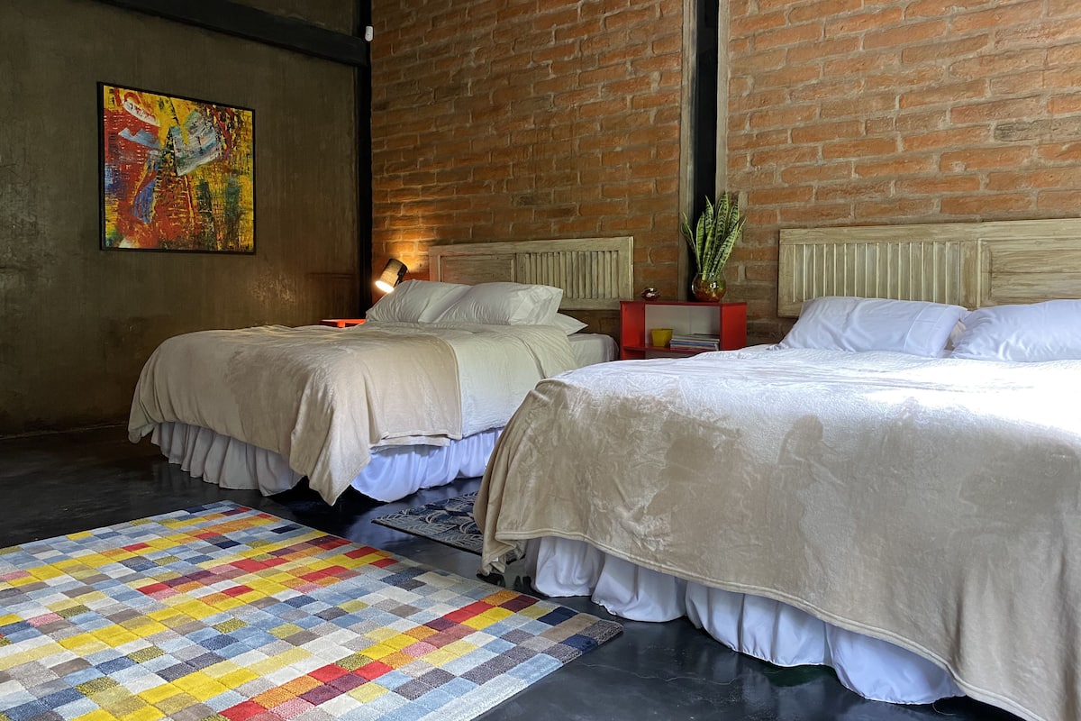 Habitación de hotel con dos camas, un tapete colorido, una pared de ladrillo y un cuadro.