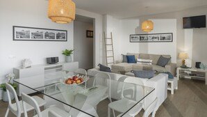 A modern living room with a glass dining table, white sofas, and a TV mounted on the wall.