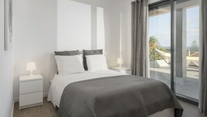 A modern bedroom with a large bed, white nightstands, and a view of a balcony with outdoor furniture.