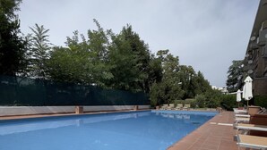 A swimming pool with tiled edges, surrounded by lounge chairs and umbrellas, with trees and a building in the background. - A stone's throw from San Pietro and Villa Pamphili (Roma)