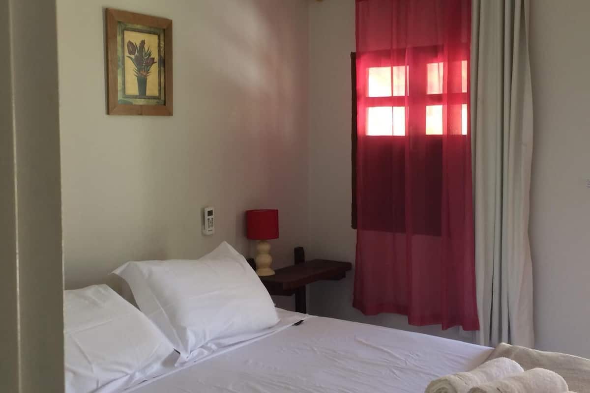 A hotel room with a bed, a bedside table with a lamp, a window with red curtains, and a framed picture on the wall.