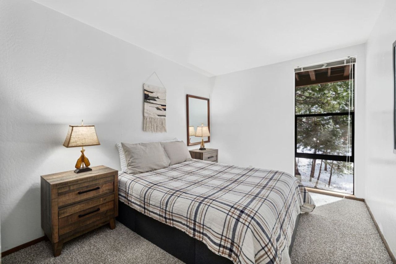 Un dormitorio con cama, dos mesitas de noche, lámpara, espejo y ventana con vista a árboles.