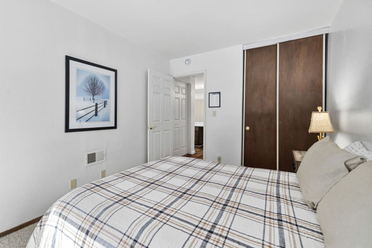 A bedroom with a bed, a door, a framed picture, a lamp, and a wall-mounted light switch.