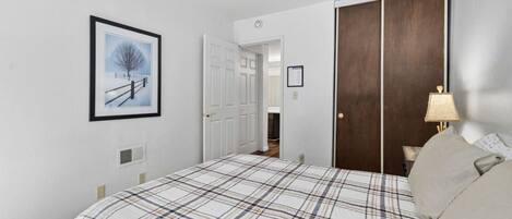 A bedroom with a bed, a door, a framed picture, a lamp, and a wall-mounted light switch.