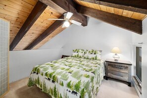 Ein Schlafzimmer mit schrägverdeckter Decke, einem Bett mit Waldmotiv, einem Nachttisch mit Lampe und einem Fenster mit Blick.