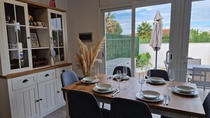 A dining area with a wooden table set for four, surrounded by chairs, and a kitchen with white cabinets and a window overlooking a garden.