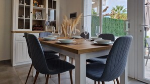 A dining area with a wooden table set for four, surrounded by dark blue chairs. A white cabinet with glass doors is to the left, displaying various items.