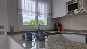 A kitchen with granite countertops, a stainless steel microwave, a window with a white curtain, and a sink with a faucet.