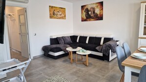A modern living room with a black and white sofa, a small round table, and a dining area with a wooden table and chairs. There are two paintings on the wall and a door leading to another room.