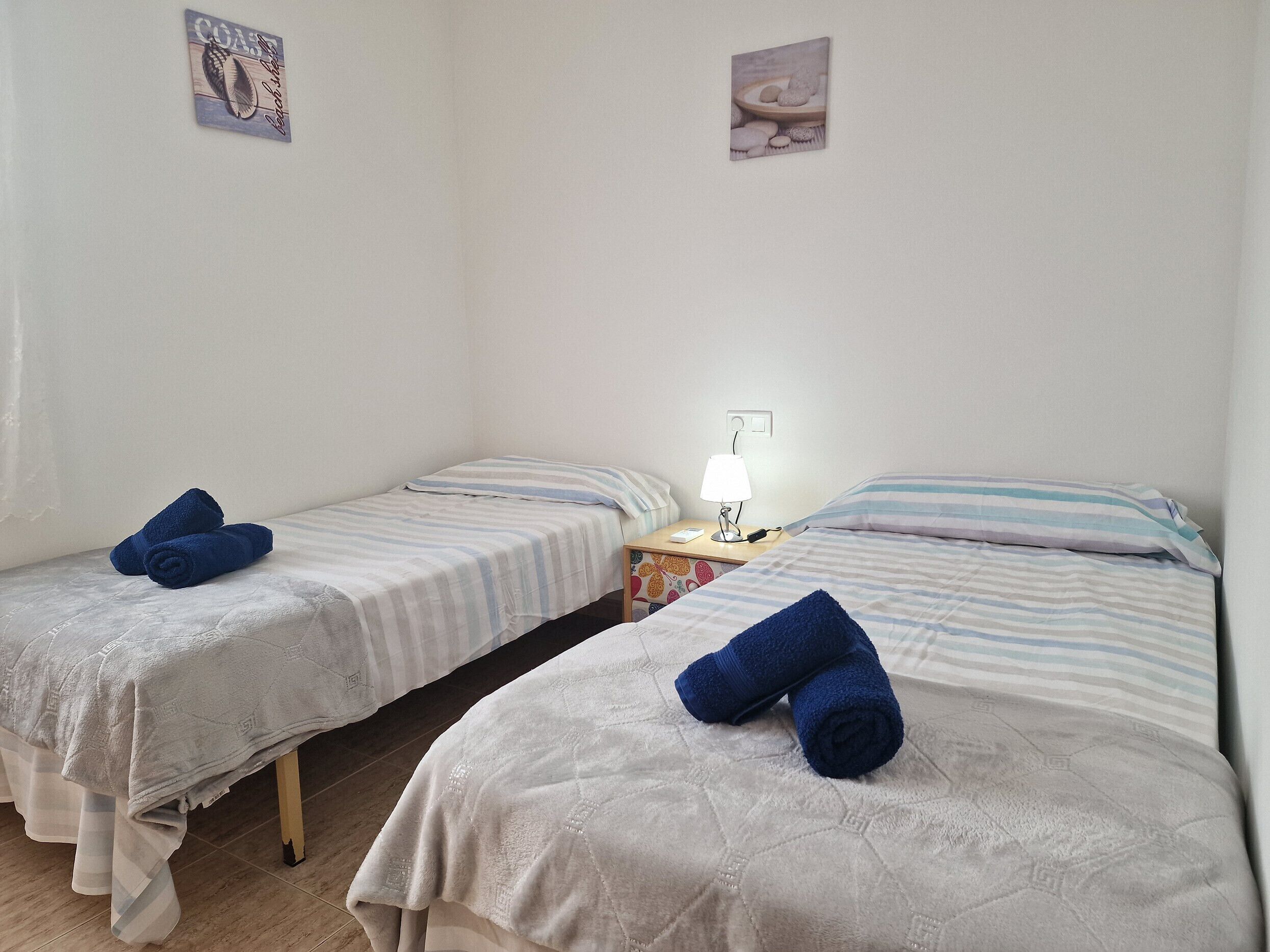 A simple room with two single beds, each with a rolled towel. There is a nightstand with a lamp and a small table with a book. The walls are plain, and there are two small pictures on the wall.