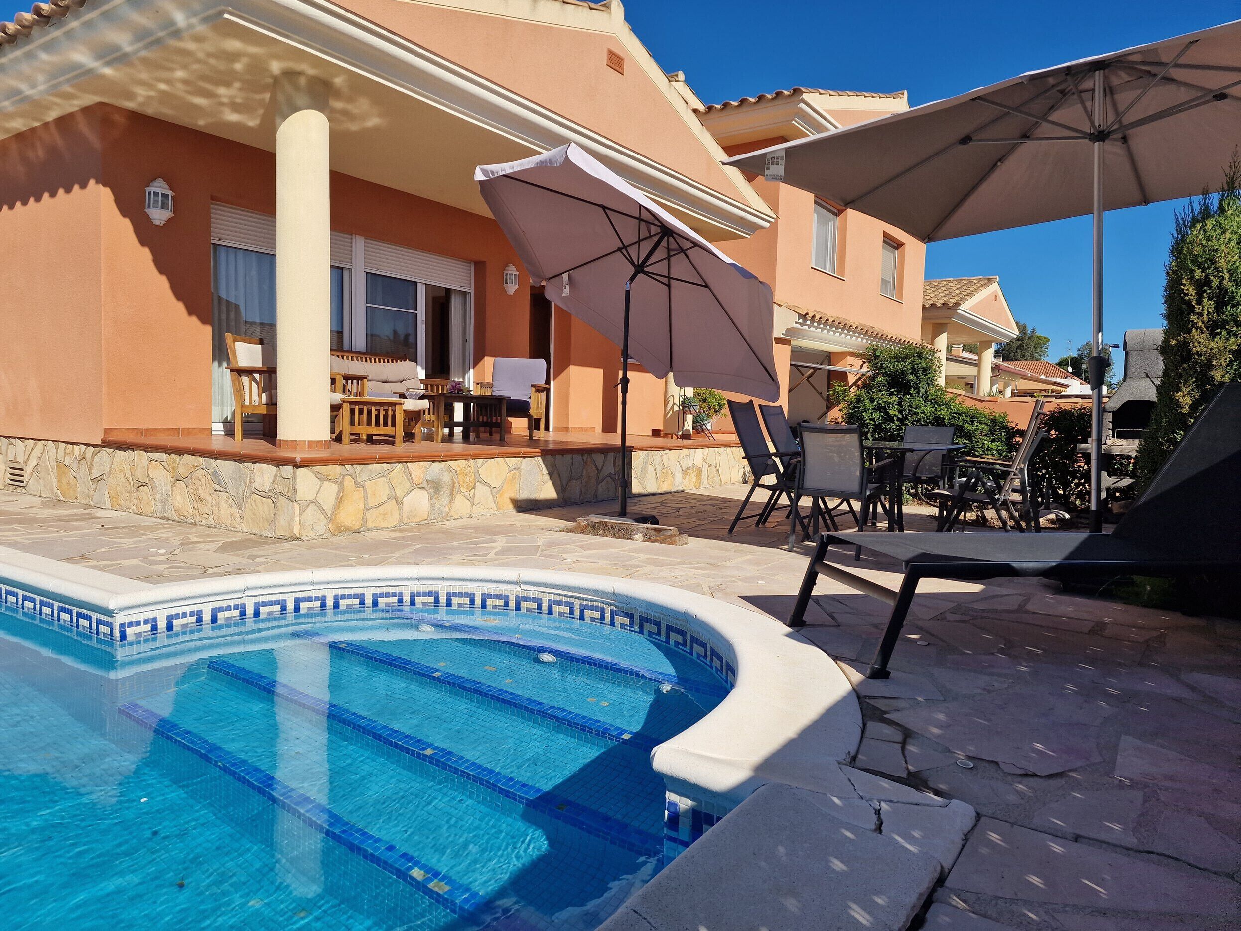 Piscina com borda azulejada, área de estar externa com guarda-sóis e uma parede de pedra.