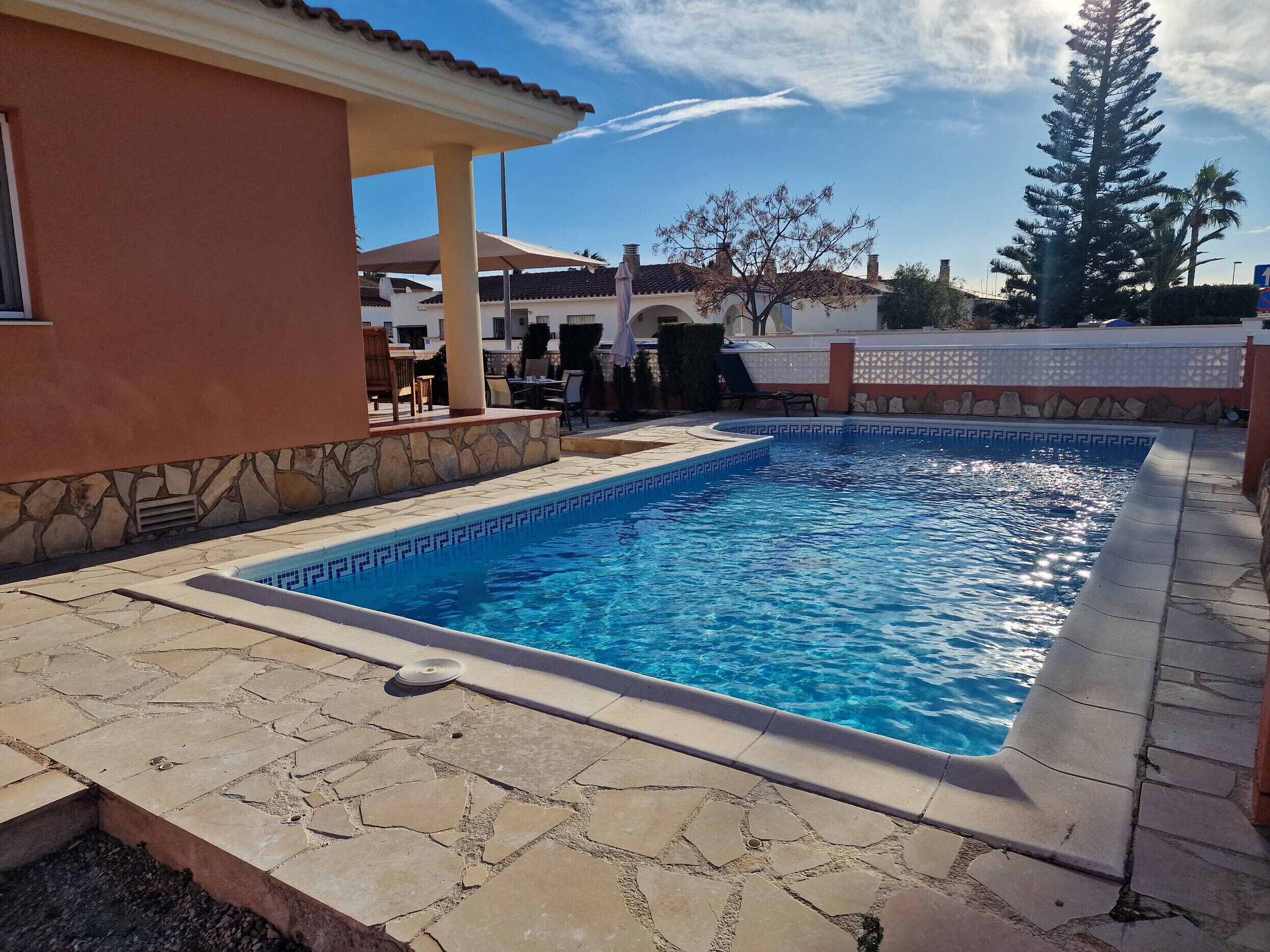 En swimmingpool med en kant av sten och en kantlist av kakel, omgiven av en uteplats med stenplattor och några solstolar.