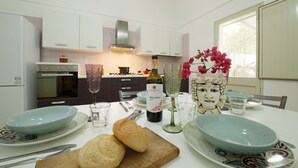 A modern kitchen with white cabinets, a stainless steel range hood, and a dining area with a table set for two. - Villa Loria - Luxurious Villa with private pool located in the city centre (San Vito Lo Capo)