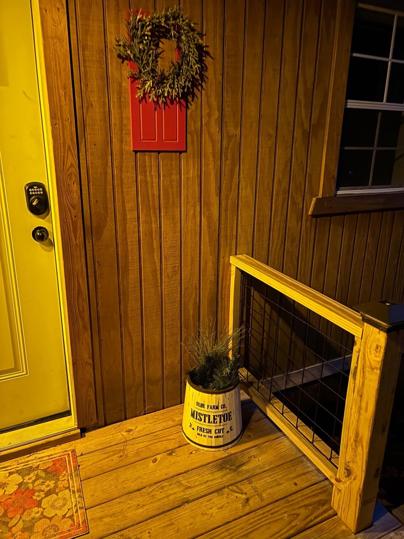 Une porte en bois ornée d’une guirlande rouge, un pot de fleurs contenant une plante étiquetée « gui », et un petit escalier en bois muni d’une rampe.