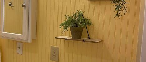 A bathroom with a toilet, sink, and mirror. There are two shelves with plants and a painting on the wall.