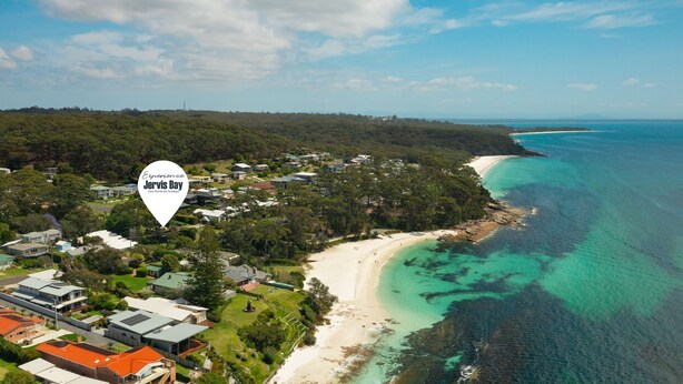 An aerial view of Jervis Bay with a coastal beach, residential area, and clear turquoise waters.