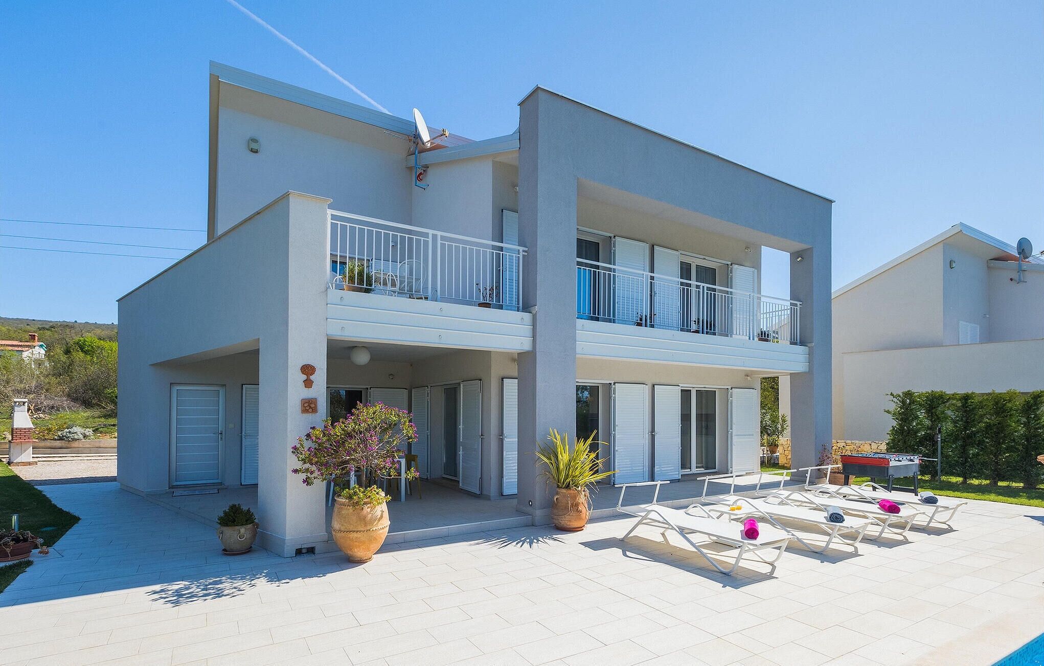 Een modern gebouw met twee verdiepingen, een plat dak, een balkon op de bovenverdieping en een overdekt terras met lounge stoelen.