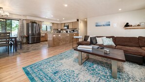 A spacious living area with a wooden floor, a brown sofa, a coffee table, and a kitchen in the background.