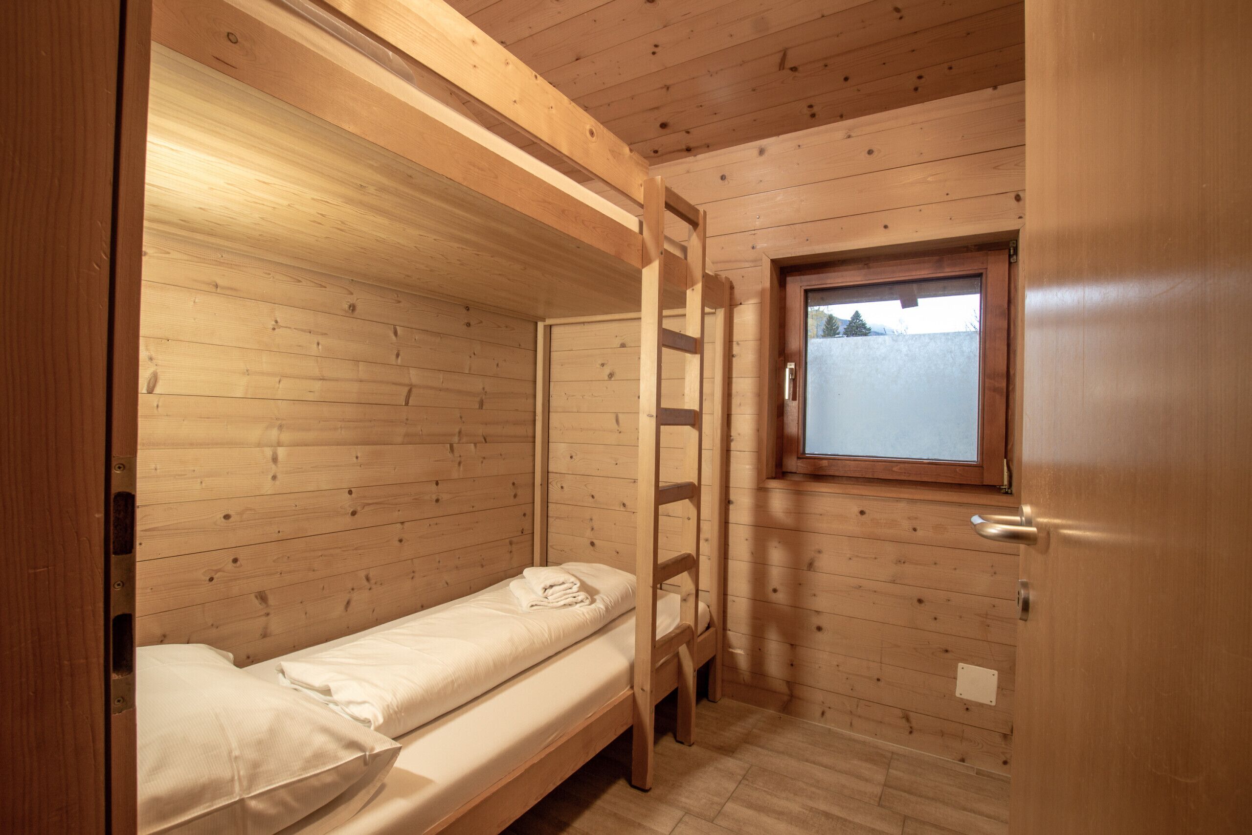 A small, wooden cabin room with bunk beds, a ladder, and a window with a view of the outside.