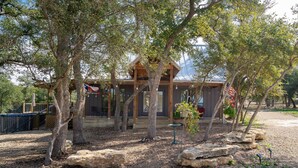 A rustic cabin with a wooden exterior, surrounded by trees and rocks, featuring a porch with a small table and potted plants.