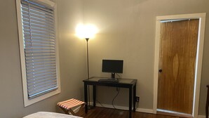 A bedroom with a bed, a desk with a computer, a lamp, a window with blinds, and a door.