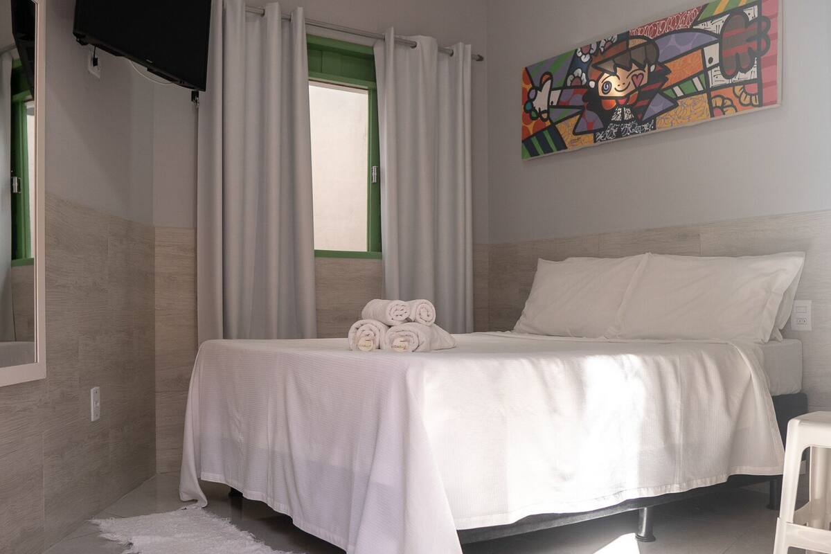 A bedroom with a bed, white curtains, a painting, and a TV mounted on the wall.