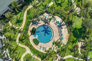 Eine Vogelperspektive zeigt einen Urlaubsort mit einem zentralen Schwimmbecken, umgeben von Liegestühlen und Palmen.