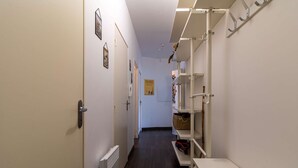 A narrow hallway with white walls, a radiator, and a door at the end. Shelves with various items are on the right wall. - Apartment near beaches in Fréhel | 4 guests | Ref 359 (FREHEL)
