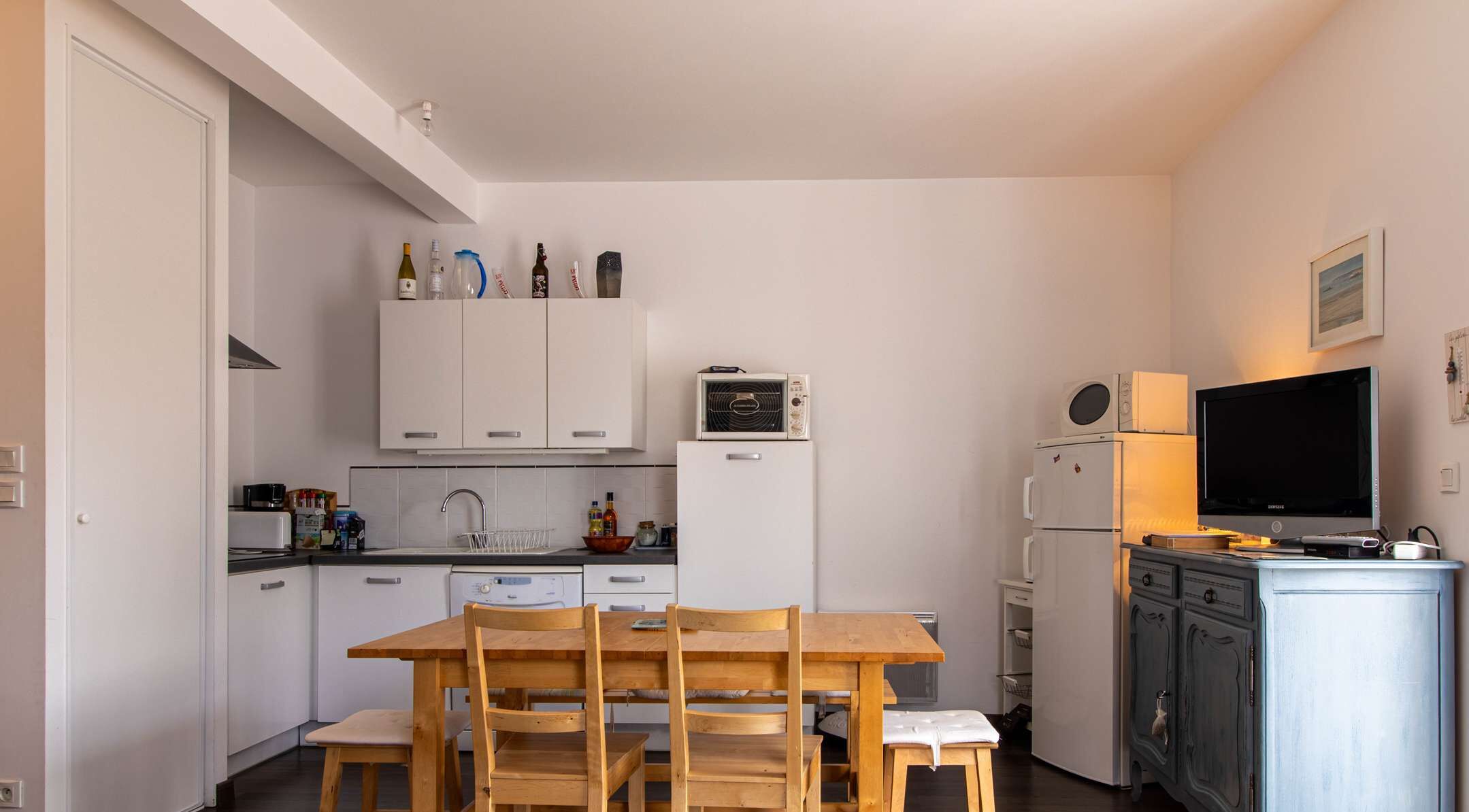 A compact kitchen with white cabinets, a wooden dining table, and a TV on a cabinet.