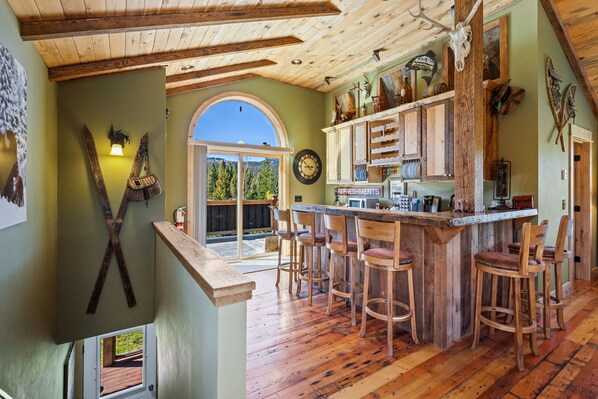 Un intérieur rustique avec des poutres apparentes, un coin bar avec des tabourets et une grande fenêtre en arc d’où l’on peut admirer la verdure.