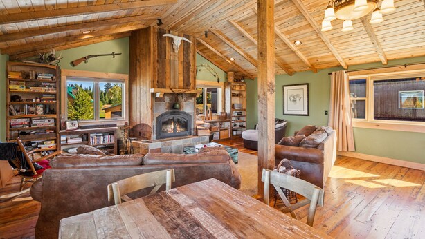 Interior rústico de una cabaña con chimenea de piedra, muebles de madera y un amplio ventanal con vistas a un bosque.