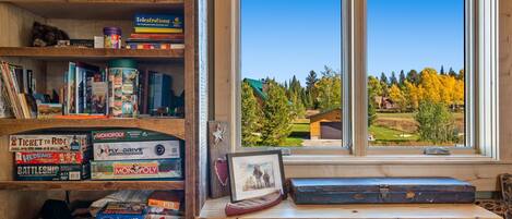 A room with a large window overlooking a scenic landscape, wooden shelves filled with board games, and a desk with a framed picture and a book.